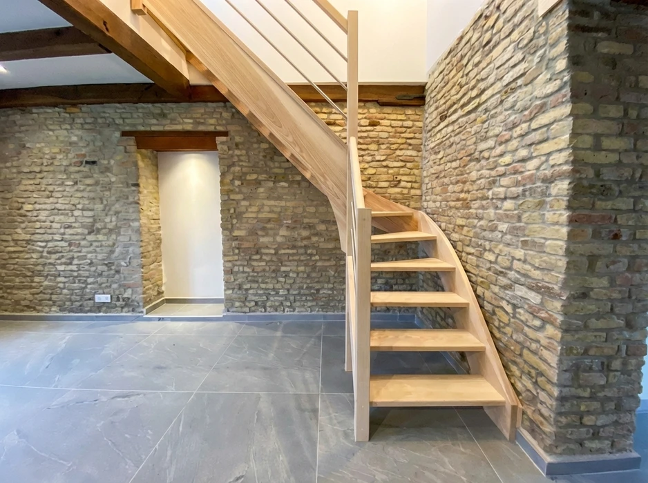 Façade en briques rouges d'un bâtiment résidentiel avec trois ouvertures : une fenêtre équipée d'un volet roulant métallique anthracite avec jardinière fleurie, une porte d'entrée en bois massif vertical naturel à arc surbaissé, et une petite porte métallique peinte noire